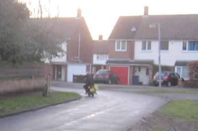 The scooter passenger is wearing a hoodie, but not a helmet, he his holding on with one hand and has his shopping in the other.